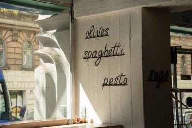 Interior wall of a cafe features handwritten words olives, spaghetti, pesto, and sugo in black script, illuminated by natural sunlight. For illustrating restaurant decor, Italian cuisine themes, or urban dining environments.