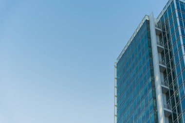 Camdan pencereleri ve çelik yapısı olan, açık mavi gökyüzüne karşı kurulmuş, modern bir ofis binası. İş, mimari veya kentsel gelişim kavramlarını resmetmek için.