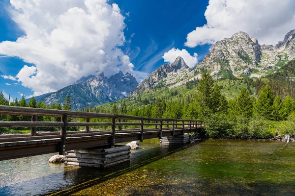 Grand Teton Ulusal Parkı 'ndaki dramatik ve fırtınalı bir gökyüzünün altında yüksek dağlara doğru bir nehri geçen bir patika köprüsü var.