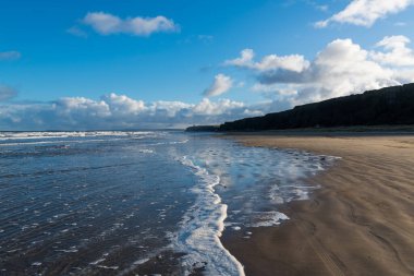 Kuzey İrlanda 'nın Benone Strand kentindeki suya yansıyan gökyüzü ve bulutlarla, dramatik bir ışık altında, kumlu bir plaja doğru yol alan araba izleri.