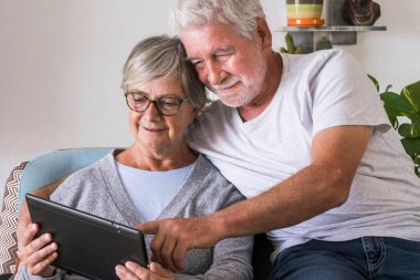 couple of seniors smiling and looking at the same tablet hugged on the sofa - indoor, at home concept - caucasians mature and retired man and woman using technology 