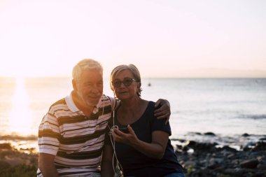 Sahilde bir çift son sınıf öğrencisi aynı telefon ve aynı şarkıyla müzik dinliyor. Gözlüklü ve emekli bir kadın yalnız eğleniyor. Arka planda deniz ve kayalar. 