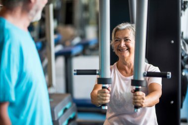 couple of two seniors at the gym training together - woman doing a exercise in a machine and the mature man is looking at her - smiling and having fun   clipart