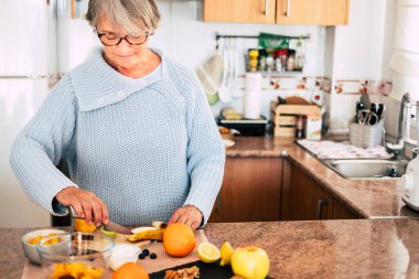 Gözlüklü büyükanne meyve kesiyor ve gülümsüyor - yaşam tarzı sağlıklı - arka planda mutfak - evde yemek pişiriyor 