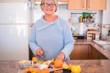Gözlüklü büyükanne meyve kesiyor ve gülümsüyor - yaşam tarzı sağlıklı - arka planda mutfak - evde yemek pişiriyor  
