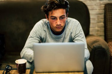teenager alone at home on the sofa with his laptop working or playing or watching videos - night with coffee and glasses on the table  clipart