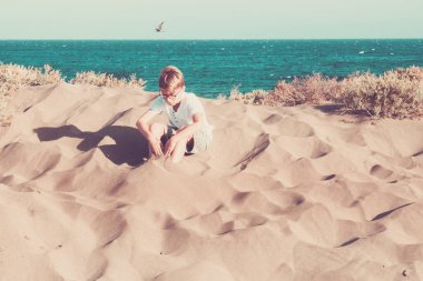 Kumsalda kumla tek başına oynayan genç bir çocuk. Gezgin ve tatil hayatı tarzı. Gözlüklü beyaz bir çocuk, deniz dibinde oturuyor. 