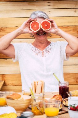 mature and retired senior at home playing and joking with two orange on her eyes with a breakfast on the table - 60s showing at the camera she joking and having fun  clipart