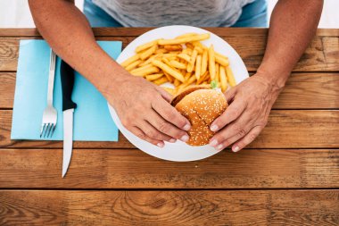 Ahşap bir masada elinde cipslerle hamburger tutan olgun bir kadın. Fast food ve sağlıksız bir yaşam tarzı.  