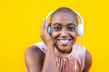 Yaklaş ve kameraya bakıp kulaklıklarıyla müzik dinleyen Afro-Amerikan bir kadının portresini yap. Sarı arka plan. 