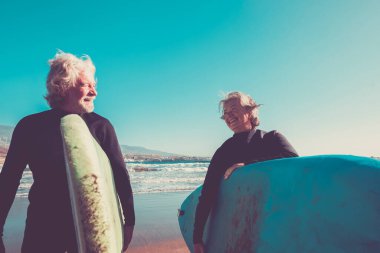 couple of seniors at the beach with black wetsuits holding a surftable ready to go surfing a the beach - active mature and retired people doing happy activity together in their vacations or free time  clipart