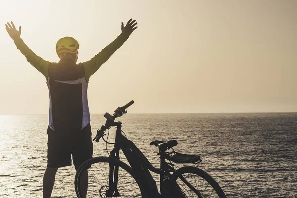 man in front of sea with arms up looking the horizon at the sunset with his bike - freedom lifestyle concept and rider senior
