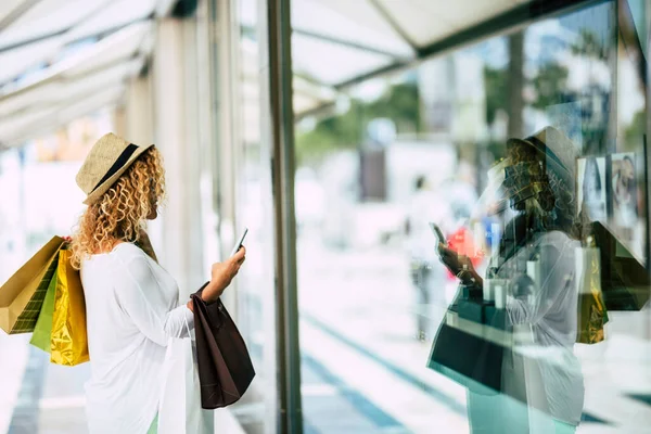 one woman alone and isolated on a mall looking at the glass of a store at the city with her phone on the hand - trendy woman want to buy something and with three bags on her back