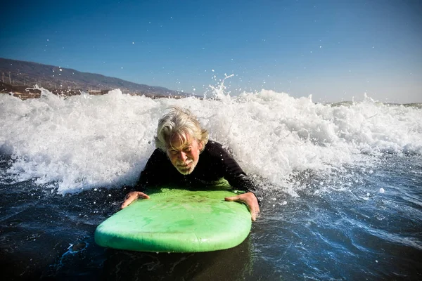 先輩は黒のウェットスーツと緑のサーフィンで一人でビーチで海の波をサーフィンしようとしています 海での休暇とアクティブな引退した男 — ストック写真
