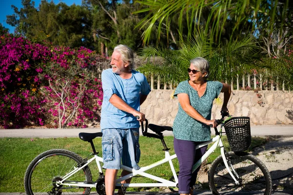 beautiful couple of senior and mature married, riding a double bike, tandem, together to have fun with a great and sunny day - woman and man - standing looking at the back