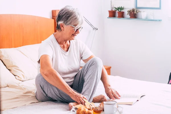 stock image mature woman sitting on the bed reading a book and eating croissant and crackers and drinking juice of orange - retired senior having breakfast isolated and alone at the morning  