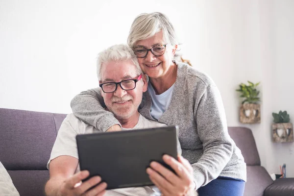beautiful couple of seniors or mature people at home hugged looking at the same tablet or laptop - retires seniors with glasses using the new technology