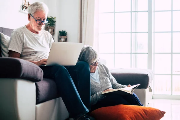 stock image couple of seniors at home - man with laptop and glasses sitting at the sofa - woman sitting on the ground reading a book - indoor and love concept 