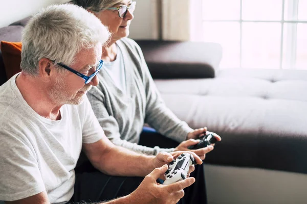 couple of caucasian retired seniors playing video games together at home sitting on the sofa - mature woman and man married - two get engaged forever - holding a controller or a joystick
