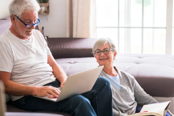 stock image couple of seniors at home - man with laptop and glasses sitting at the sofa - woman sitting on the ground reading a book - indoor and love concept 