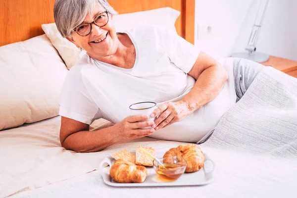 stock image mature woman sitting on the bed eating croissant and crackers and drinking juice of orange - retired senior having breakfast isolated and alone at the morning 