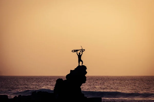 adult holding on his arms up a bike at the beach on a big rock - freedom concept and lifestyle - great sunset - ocean and sea with waves at the background