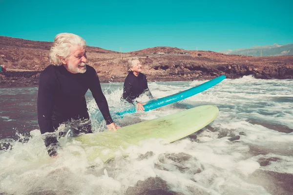 2人の高齢者が波に乗るか 取る方法を学ぶために水の中に入るウェットスーツやサーフボードと一緒にサーフィンに行く 夏のスポーツと健康とフィットネスライフスタイル — ストック写真