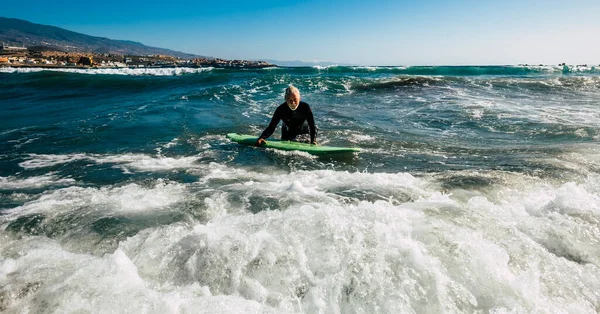 先輩は黒のウェットスーツと緑のサーフィンで一人でビーチで海の波をサーフィンしようとしています 海での休暇とアクティブな引退した男 — ストック写真