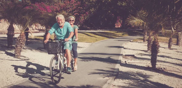 stock image beautiful couple of seniors riding a tandem together having fun in a great park with plants and palms - happy two mature people riding a bike and laughing  