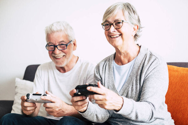 couple of seniors sitting on the sofa and playing video games holding a controller - people have fun and enjoy  together laughing - indoor and at home concept 