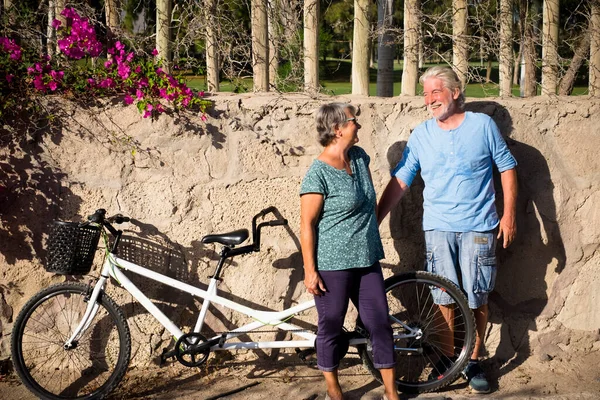 couple of two seniors and mature people standing with the wall at the background in a park with their tandem or double bike smiling and talking