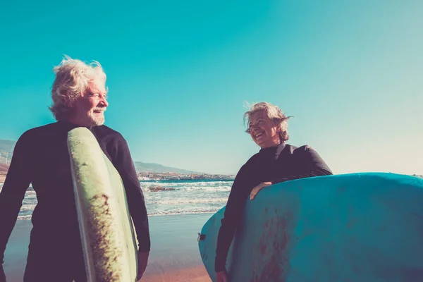 ビーチで黒のウェットスーツを着た高齢者のカップルは ビーチをサーフィンする準備ができているサーフィンをする準備ができています アクティブな成熟し 引退した人々は休暇や自由な時間で一緒に幸せな活動をしています — ストック写真