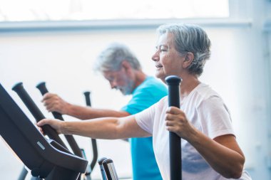 woman using a precor at the gym - active senior with his man at the background - couple of pensioners doing exercises together  clipart