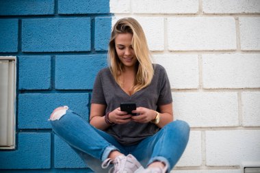 Güzel ve sarışın bir kadın telefonuyla oturuyor mavi-beyaz bir duvar ve arka plan - telefona bakan bir genç.  