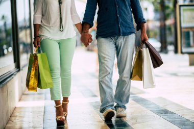 İki yetişkinin bacakları üzerinde giysi olan beş alışveriş çantasıyla alışveriş merkezinde yürürken. Kadın ve erkeğin ilişkisi kıyafet almaya gidiyor. 