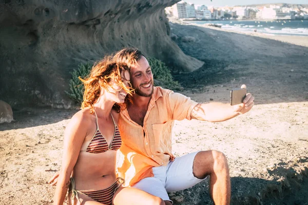 beautiful couple of adults taking a selfie together at the beach sitting on the rocks