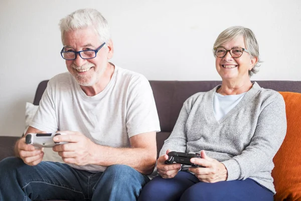 stock image couple of seniors playing video game with joystick at home - indoor enjoying and have fun with games - sitting on a sofa - smiling 
