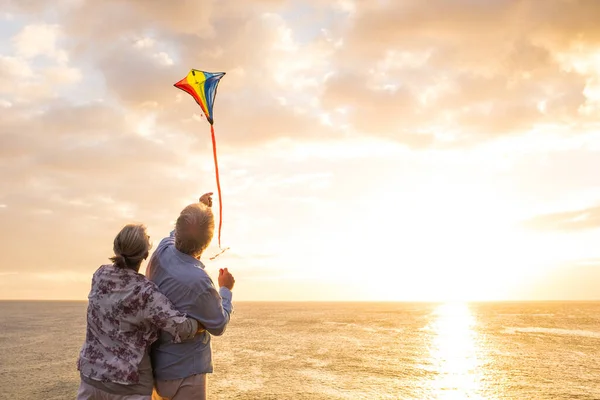 stock image close up and portrait of two old and mature people playing and enjoying with a flaying kite at the beach with the sea at the background with sunset - active seniors having fun  