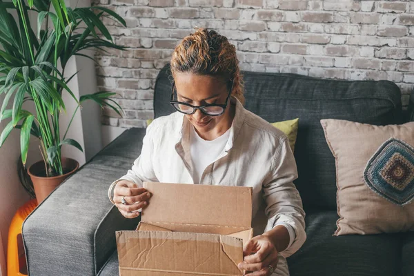 Woman unpacking delivery box opening package at home. Happy young lady looking at carton box while sitting on sofa in living room at modern apartment. Caucasian female checking out delivered stuff.