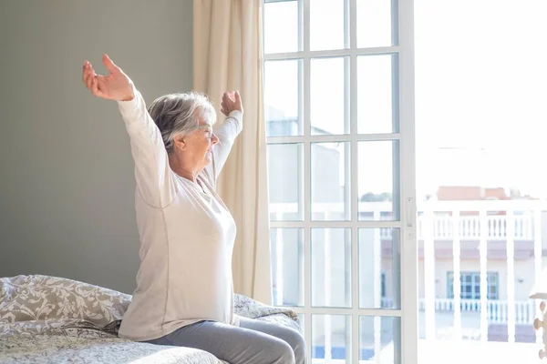 Senior woman sitting on bed with arms wide outstretched and eyes closed in bedroom next to window at her apartment. Satisfied old woman stretching hands on bed at home