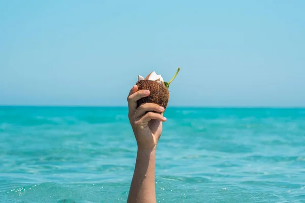 Close up of hand of an unrecognizable person holding half coconut shell with slices on coconut in it against sea and sky. Wet hand in summer with coconut shell. Raised hand holding coconut against sea