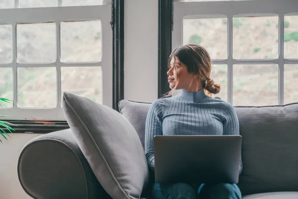 一个在沙发上使用笔记本电脑的年轻女人看着窗户 想着未来和工作 女性有识之士放松工作或上网 — 图库照片