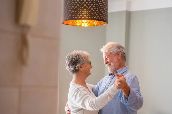 Cheerful senior couple dancing and smiling looking at each other. Elderly happy couple celebrating by dancing in living room. Romantic old aged couple holding hands and dancing together at home