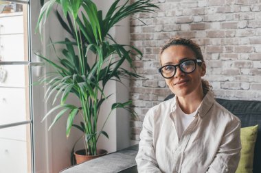 Rahat giysiler içinde rahat bir genç kadının portresi rahat bir modern oturma odasında kanepede oturmuş kameraya bakıyor ve gülümsüyor. Kafkasyalı mutlu bir kadın dairesinin kanepesinde dinleniyor. 