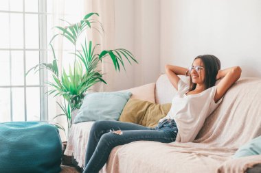 Gözlüklü mutlu genç bir kadın evinin oturma odasında elleri başının arkasında kanepede dinleniyor. Tasasız ve memnun bir bayan yırtık kotla modern apartmanda koltukta dinleniyor. 