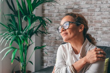 Düşünceli, rahat, gündelik giysiler içinde genç bir kadın rahat modern oturma odasında koltukta oturuyor ve başka tarafa bakıyor ve gülümsüyor. Kafkasyalı kadın dairesinin kanepesinde dinleniyor. 