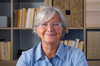 Gözlüklü, gri saçlı, kitaplığın önündeki kameraya bakan son sınıf öğrencisi kadının portresi. Yaşlı mutlu bayan öğretmen eğitim dersi veriyor ya da video konferansı yapıyor. 