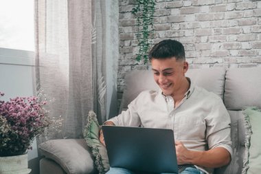 Genç mutlu bir adam internette dizüstü bilgisayarıyla geziniyor. Kanepede oturuyor ve boş zamanın tadını çıkarıyor ve yalnız başına dinleniyor. Girişimci çocuk çalışıyor ve geleceğini inşa ediyor.. 