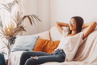 Gözlüklü mutlu genç bir kadın evinin oturma odasında elleri başının arkasında kanepede dinleniyor. Tasasız ve memnun bir bayan yırtık kotla modern apartmanda koltukta dinleniyor. 