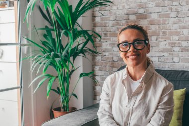 Rahat giysiler içinde rahat bir genç kadının portresi rahat bir modern oturma odasında kanepede oturmuş kameraya bakıyor ve gülümsüyor. Kafkasyalı mutlu bir kadın dairesinin kanepesinde dinleniyor. 